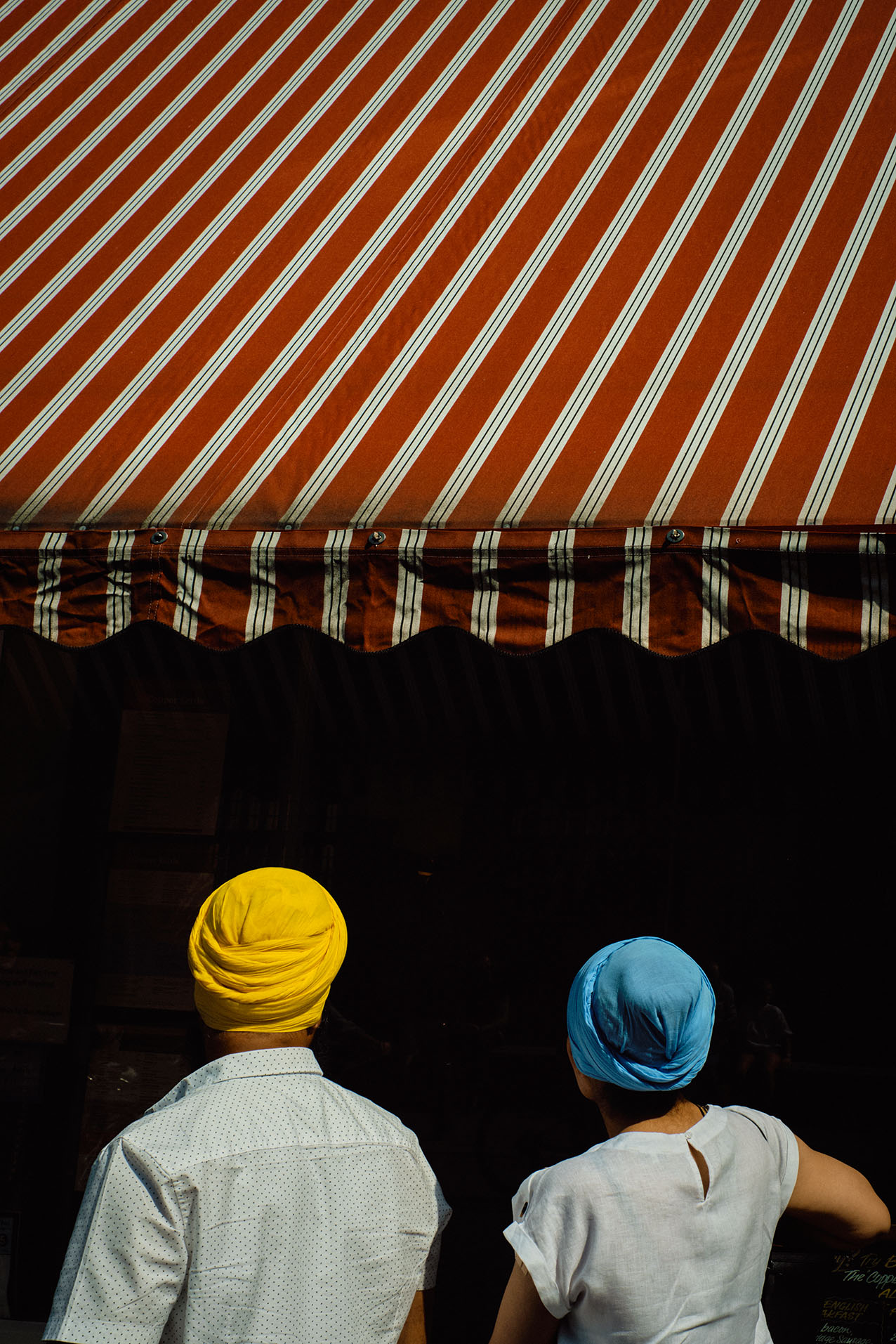 A man in a flat cap looks into the raised white bonnet of a classic car outside a green cafe